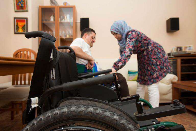 Primer pla d'una cadira de rodes i al fons una persona amb discapacitat rebent suport de la seva assistent personal