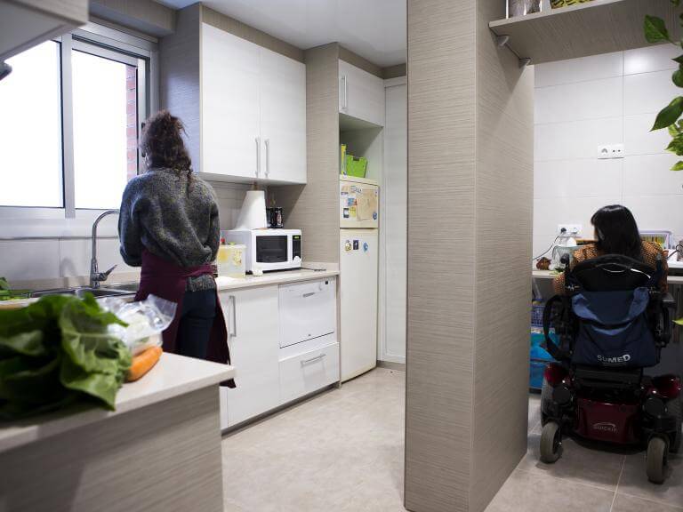 Dos mujeres de espaldas (una de ellas usuaria de silla de ruedas) en una cocina preparando algo de comida sobre los mármoles