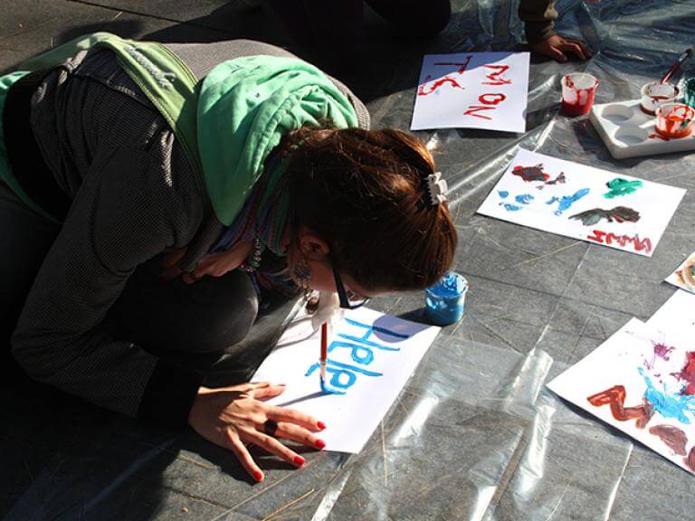 Una chica de rodillas en el suelo intentando escribir sobre un papel su nombre con un pincel puesta en la boca. 