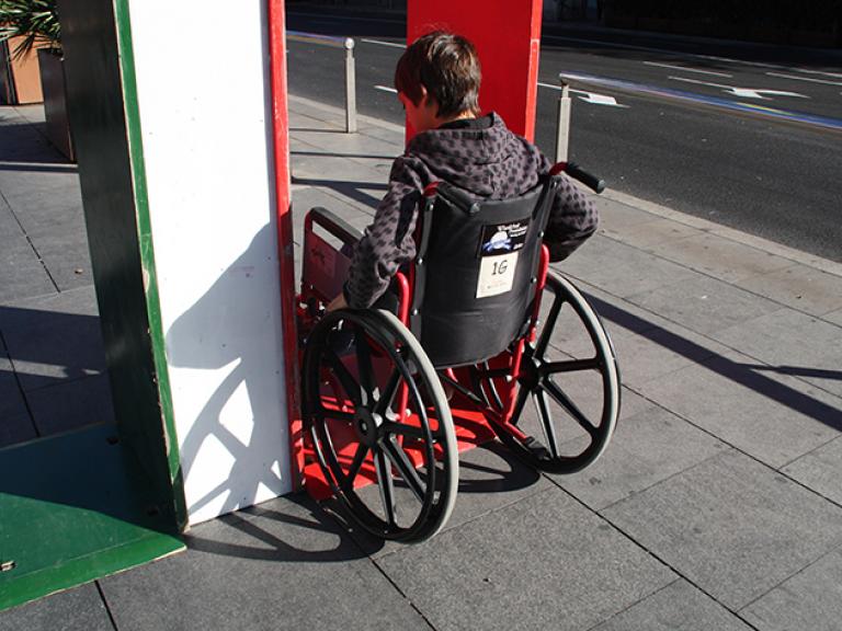 Un noi assegut en una cadira de rodes intentant passar per una estructura que simula l'entrada d'una porta. La imatge mostra dues opcions d'entrada (o porta) una pintada de verd, que és accessible, i una de vermella (que és massa estreta, tot i que permet el pas).