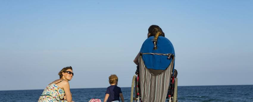 Chica usuaria de silla de ruedas en la playa (de espaldas mirando al mar) acompañada de otra chica y una niña pequeña