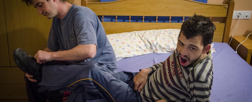 Noi amb discapacitat al seu llit que està sent ajudat per un altre noi que li està posant les sabates