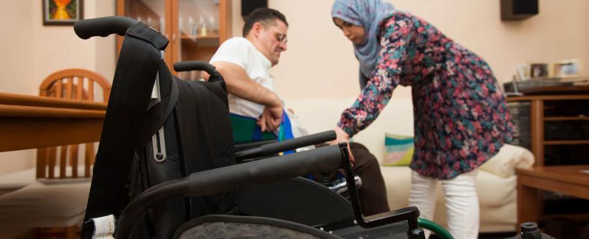 Primer plano de una silla de ruedas y al fondo una persona con discapacidad recibiendo apoyo de su asistenta personal