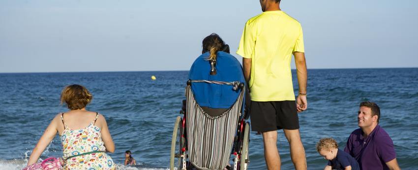 Una persona usuaria de silla de ruedas en la playa, acompañada por su asistente personal y otras personas