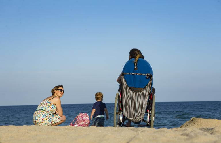 Noia usuària de cadira de rodes a la platja  (d'esquenes mirant el mar) acompanyada d'una altra noia i un infant petit
