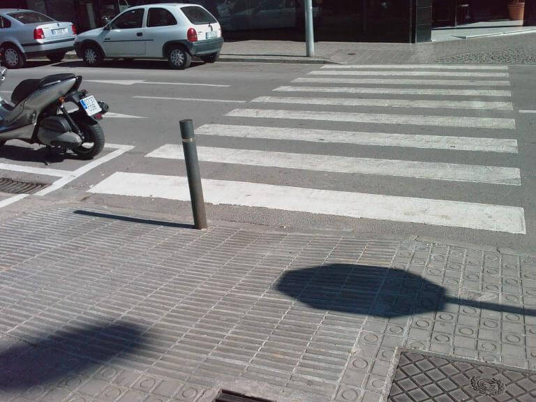 Imatge d'un carrer no accessible. S'hi veu un pas de zebra en l'inici del qual hi ha al mig un pal per evitar que els vehicles es fiquin en la vorera