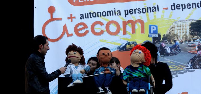 Escenario con una actuación de los títeres de ECOM con un fondo de escenario que es una pancarta reivindicativa en el Día Internacional de la Discapacidad