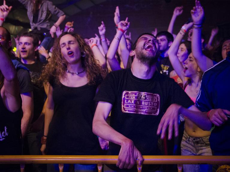 Gent jove ballant i amb els braços alçats, rera una tanca de seguretat, gaudint d'un concert. En primera fila un jove amb paràlisi cerebral