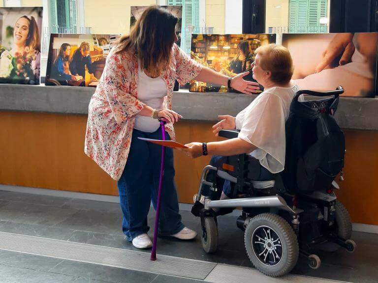 Dues dones amb discapacitat (una usuària de cadira de rodes i l'altra amb una crossa a la mà) davant d'un finestral i de diverses fotografies de dones amb discapacitat, de l'exposició 'Dones diverses' d'ECOM