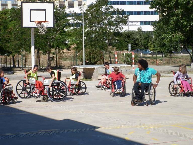 Alumnes al pati d'una escola asseguts a cadires de rodes i atenent les instruccions d'un noi amb discapacitat (també usuari de cadira de rodes) sobre com ho han de fer per moure’s en la cadira