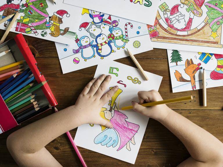 Las manos de una niña haciendo dibujos sobre una mesa llena de dibujos y colores